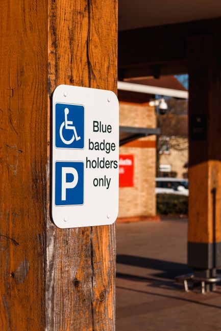 A close-up image of a yellow wheelchair accessible parking symbol painted on a dark asphalt surface, with visible texture and small gravel particles. The painted lines are slightly worn and weathered,
