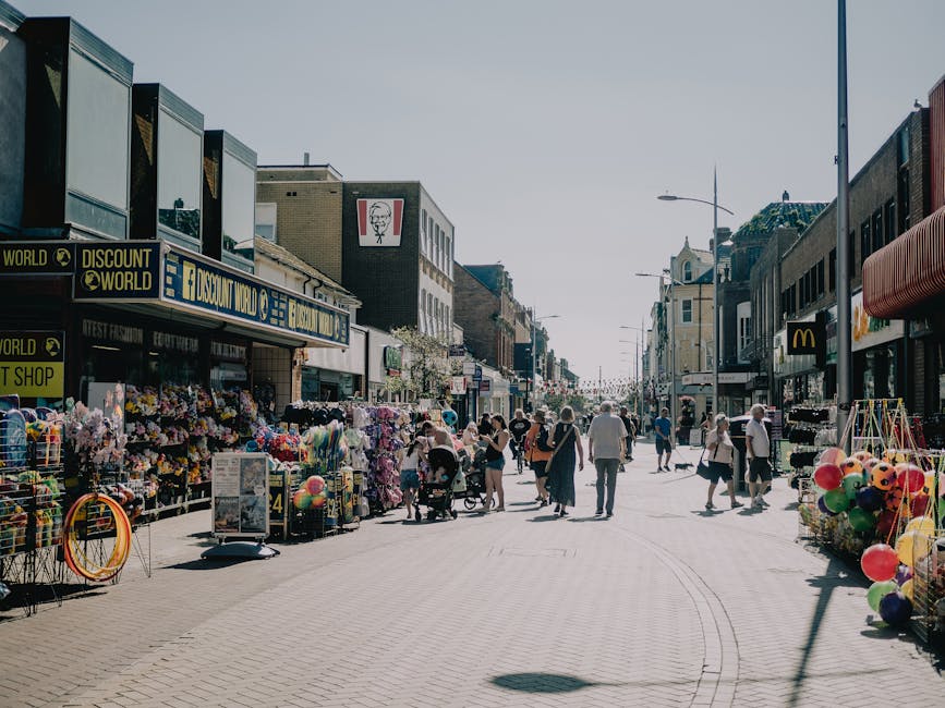 A busy pedestrian street during daylight with various shops on both sides, including a discount store with a blue and yellow sign, and a McDonald's outlet. The street is filled with people walking, some pushing strollers or carrying shopping bags. Outside the shops, there are displays of colourful toys, balloons, and flowers, with some items arranged on racks and tables. The pavement is light-coloured and appears to be concrete, with subtle tire tracks likely from a moving vehicle. On the right side, a service van from Man with Van Redbridge is partially visible, parked near the sidewalk. The scene suggests an active shopping area, possibly in the context of house removals or furniture transport, with ambient natural light indicating a clear, sunny day. Building facades consist of brick and other materials, typical of urban commercial districts in the UK, fitting with the theme of relocation or home moving services in the photo's context.