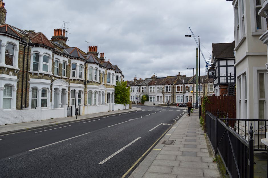 A quiet residential street on an overcast day with a row of Victorian-style terraced houses on the left, featuring white facades, bay windows, and red-tiled roofs with brick chimneys. The street has a smooth asphalt surface marked with lane lines, and a paved sidewalk on the right side with a black metal railing and some potted plants. In the foreground, there are no visible vehicles or furniture, but cranes and construction sites can be seen in the background, indicating ongoing development. The area is illuminated by tall modern streetlights, with one person walking along the sidewalk. The scene depicts an urban environment suitable for house removals or furniture transport, with a focus on the peaceful street setting that [COMPANY_NAME] may assist with for home relocation or moving services related to the Gants Hill to Ilford route.