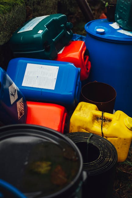 A collection of brightly coloured plastic containers, including blue, red, yellow, and black, are stacked on top of each other outdoors, some with labels or lids visible. These containers are placed on the ground near a dark garden area, with a portion of a black garden hose visible in the foreground. The scene appears to be part of a home relocation process, where packing materials and equipment are prepared for transport or disposal. The containers are made of durable plastic, some sealed with clips or screws, and are likely used for storing or transporting liquids or small items. A man with van Redbridge would manage the loading of such items into a van as part of efficient furniture transport and household moving services, ensuring safe handling and organization during the packing and moving process.