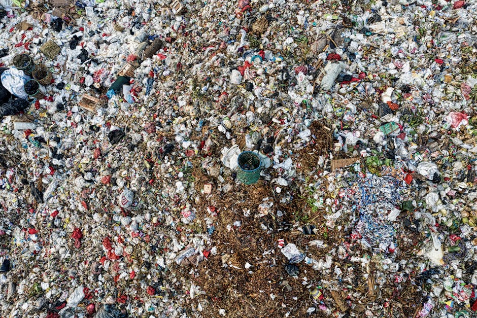 A vast outdoor area covered with a thick layer of mixed household waste, including plastic bottles, paper, cardboard, food packaging, and small scraps of fabric. Among the debris, a few larger items such as a blue plastic bucket, wooden pallets, and scattered bags are visible. The waste appears to be spread across a ground surface composed of dirt and small stones, with some areas slightly disturbed by shoveling or movement. The scene likely represents a waste disposal site or landfill associated with the final stages of home relocation or disposing of household materials before a move. The lighting is natural, suggesting daytime conditions. Man with Van Redbridge is aware of the importance of proper waste disposal in house removals, ensuring safe and compliant clearance services as part of their comprehensive moving logistics, which may include waste management and hazardous waste guidance for Redbridge moves.