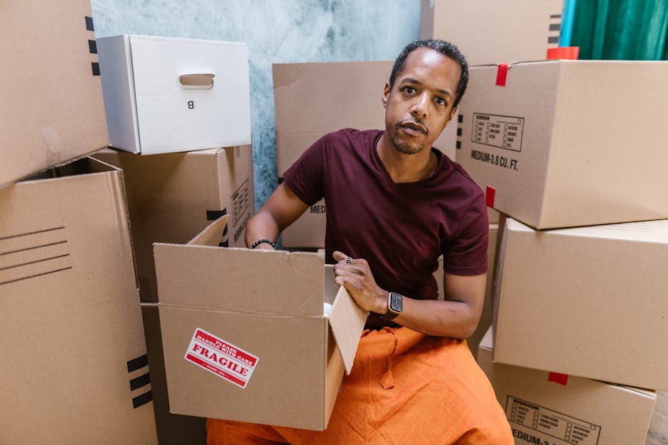 A man with dark curly hair and a serious expression, wearing a maroon t-shirt and orange pants, is positioned inside a room surrounded by unpacked cardboard boxes, some labeled with red 'Fragile' stic