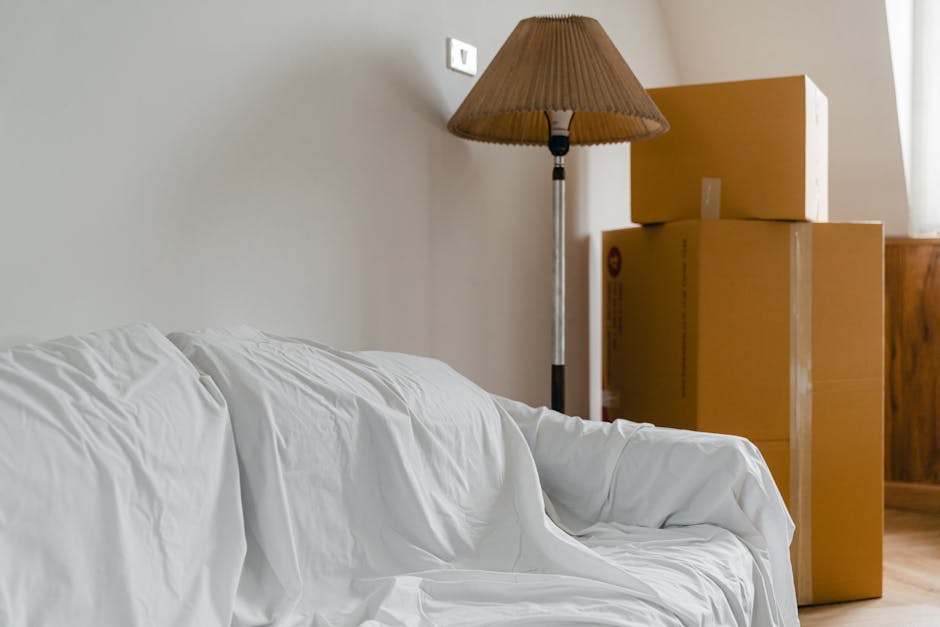 A man with dark curly hair and a serious expression, wearing a maroon t-shirt and orange pants, is positioned inside a room surrounded by unpacked cardboard boxes, some labeled with red 'Fragile' stic