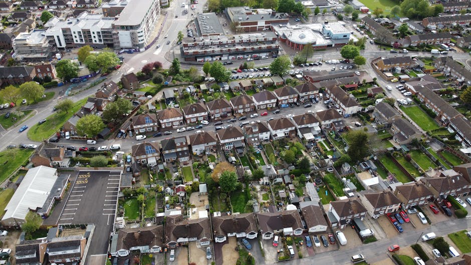 An aerial view of a residential neighbourhood showing a row of terraced houses with small private gardens, some with lawn areas and flower beds. Many houses have cars parked in driveways and along the street. A large commercial building with a flat roof and a nearby parking lot are visible in the background, alongside wider roads and green spaces with trees. The image captures the typical setting for house removals or moving logistics in Redbridge, with clear access points for loading furniture and packing materials from homes to moving vans. The outdoor scene is well-lit, indicating daytime, and the overall environment suggests a quiet, suburban area suitable for household relocation activities, with some visible equipment like wheelie bins and garden sheds complementing the scene. Man with Van Redbridge operates in such environments for efficient, professional removals services.