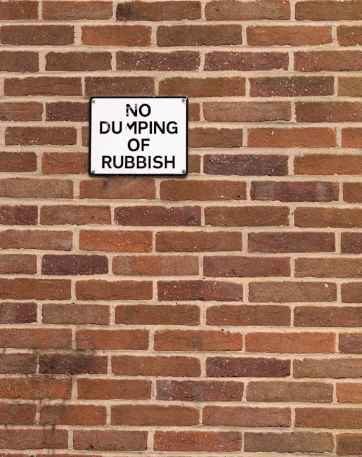 A close-up image of a rectangular, white metal sign mounted on a red brick wall. The sign displays the black, capitalized text 'NO DUMPING OF RUBBISH' with some letters slightly faded or obscured. The brick wall behind the sign features horizontal rows of reddish-brown bricks with light gray mortar, creating a textured background. The lighting is natural, with no shadows or reflections visible. The sign's placement suggests it is intended as a warning related to waste disposal and is situated in an outdoor area likely near a residential or commercial building. The image emphasizes the importance of maintaining cleanliness and adhering to local rules, aligning with the context of house removals or relocation logistics handled by Man with Van Redbridge, especially when considering moving regulations such as skip-permit rules for safe and compliant home relocations.