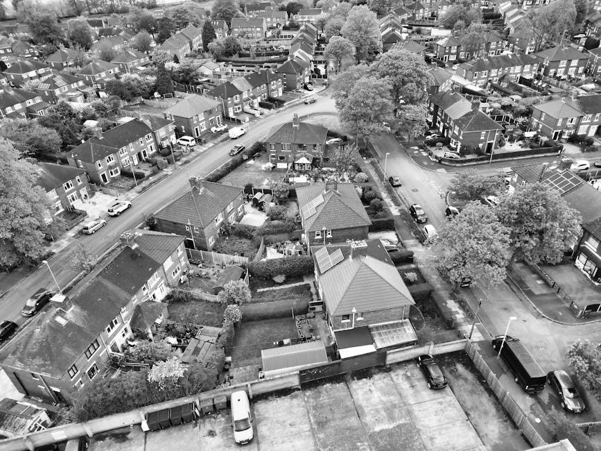 An aerial view of a residential neighbourhood showing a row of terraced houses with small private gardens, some with lawn areas and flower beds. Many houses have cars parked in driveways and along the street. A large commercial building with a flat roof and a nearby parking lot are visible in the background, alongside wider roads and green spaces with trees. The image captures the typical setting for house removals or moving logistics in Redbridge, with clear access points for loading furniture and packing materials from homes to moving vans. The outdoor scene is well-lit, indicating daytime, and the overall environment suggests a quiet, suburban area suitable for household relocation activities, with some visible equipment like wheelie bins and garden sheds complementing the scene. Man with Van Redbridge operates in such environments for efficient, professional removals services.