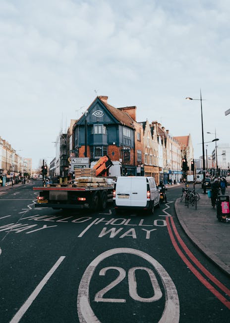 A white flatbed van is parked on a busy urban street in the process of a home relocation, with large wooden furniture pieces wrapped in protective packaging and secured with straps loaded onto the vehicle. The van is positioned near the curb on a road marked with a 20 mph speed limit and directional lines. Behind the van, there is a black moving truck with open back doors and a stack of cardboard boxes and furniture wrapped in plastic on its loading platform. In the background, multi-story brick and painted commercial buildings line both sides of the street, with pedestrians walking on the pavement, some near bicycles parked along the sidewalk. Streetlights, traffic signals, and clear signs are visible under a partly cloudy sky, indicating a typical urban setting suitable for reliable furniture transport and packing during a house removal or relocation process. The scene is representative of professional moving logistics, often handled by companies such as Man with Van Redbridge, supporting efficient loading times and optimal van routes for local house removals.