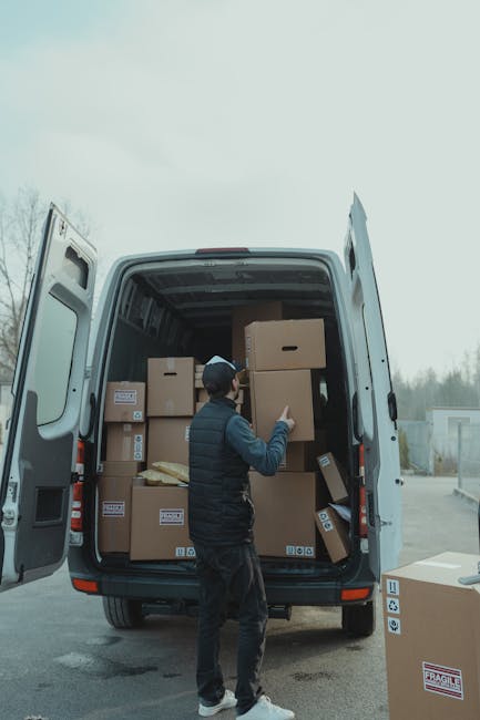A person dressed in a blue and black jacket is standing next to a moving van with its rear doors open, filled with cardboard boxes of various sizes, some sealed with packing tape and others with visible flaps, arranged on the van's floor. The individual is holding a clipboard and appears to be checking or noting details during a home relocation process. A hand truck or trolley with a black handle and metal frame, loaded with stacked boxes, is positioned next to the person, ready for transport. The area is outdoors, with the van parked on a paved surface, and the surroundings suggest a residential or commercial property entrance. This scene reflects a professional furniture transport or packing and moving operation, typical of house removals services such as those offered by Man with Van Redbridge, assisting with efficient loading and transport logistics during a house move near Seven Kings (IG3).