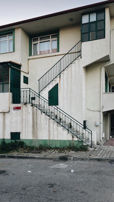 The exterior of a multi-storey residential building with a white façade, featuring a metal staircase with black railings leading from the ground level to an upper floor. The staircase is positioned ad