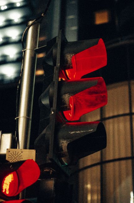 Close-up view of a traffic light at night showing a red signal, mounted on a metal pole with a silver finish. The traffic light is positioned next to a building with horizontal metal siding illuminated by ambient lighting. The red light indicates that vehicles or pedestrian crossings must stop, reflecting typical street-loading and parking regulations in Redbridge. This scene is relevant to home relocation and packing and moving processes managed by Man with Van Redbridge, highlighting the importance of adhering to local street-loading rules during moving operations. The image captures the urban environment where loading activities may be subject to specific timings and permit requirements, with the traffic light serving as a visual cue for temporary stopping during furniture transport or loading procedures in a residential or commercial area.