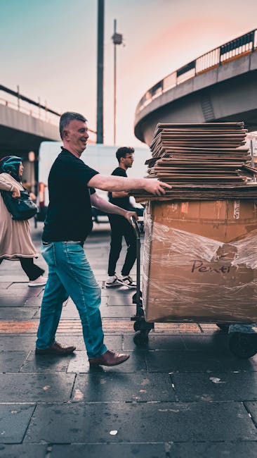 A man wearing a black top and blue jeans is pushing a large flatbed trolley loaded with numerous flattened cardboard boxes and packing materials, including plastic-wrapped items, along a paved outdoor area beneath an overpass. The trolley is positioned near a curb, suggesting a loading or unloading zone in a busy urban environment. In the background, other pedestrians walk past, and a second individual, also managing the move, can be seen nearby. The scene is captured during daytime with natural light illuminating the area, reflecting the logistics involved in a home relocation or furniture transport process. This image illustrates the typical packing and loading stage of a house removals service, with features aligned to the services offered by Man with Van Redbridge, supporting efficient and organized moving operations, especially for urgent same-day moves in Redbridge.