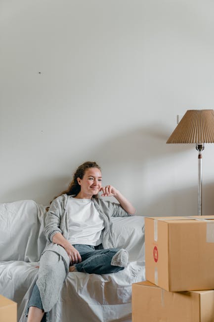 A young woman with light brown hair tied back, sitting relaxed on a white sofa inside a home, smiling and resting her chin on her hand. In front of her, there are two closed cardboard boxes, one larger and one smaller, indicating packing for a move. To the right, a tall floor lamp with a brown fabric shade provides soft lighting, while the plain light-colored wall behind features minimal decor. The scene suggests preparation for a house relocation or packing process, aligning with the services of Man with Van Redbridge, which offers residential removals and furniture transport, and illustrates the domestic environment involved in home relocation and moving logistics.