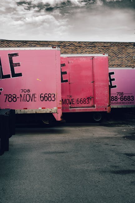 Two workers wearing yellow shirts and black gloves are seen loading large white plastic-wrapped bags onto a dark blue flatbed truck parked outdoors in a parkside area with tall trees and green foliage